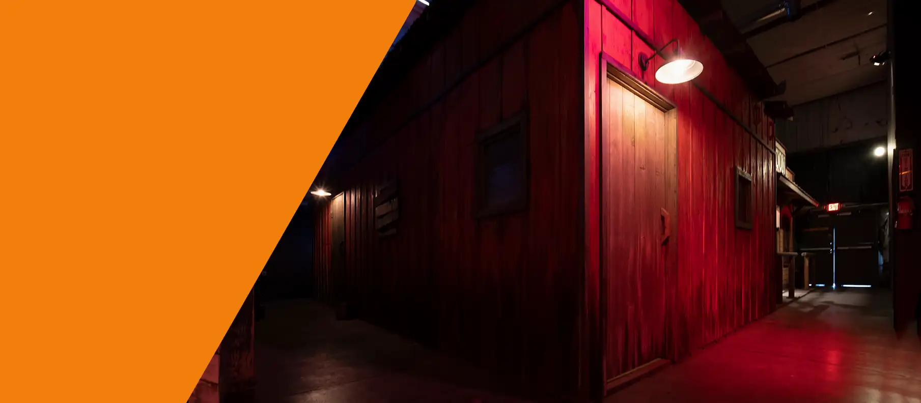 Old West town alleyway escape room setting featuring vertical red wood plank buildings, closed doors, and overhead gooseneck barn lighting.